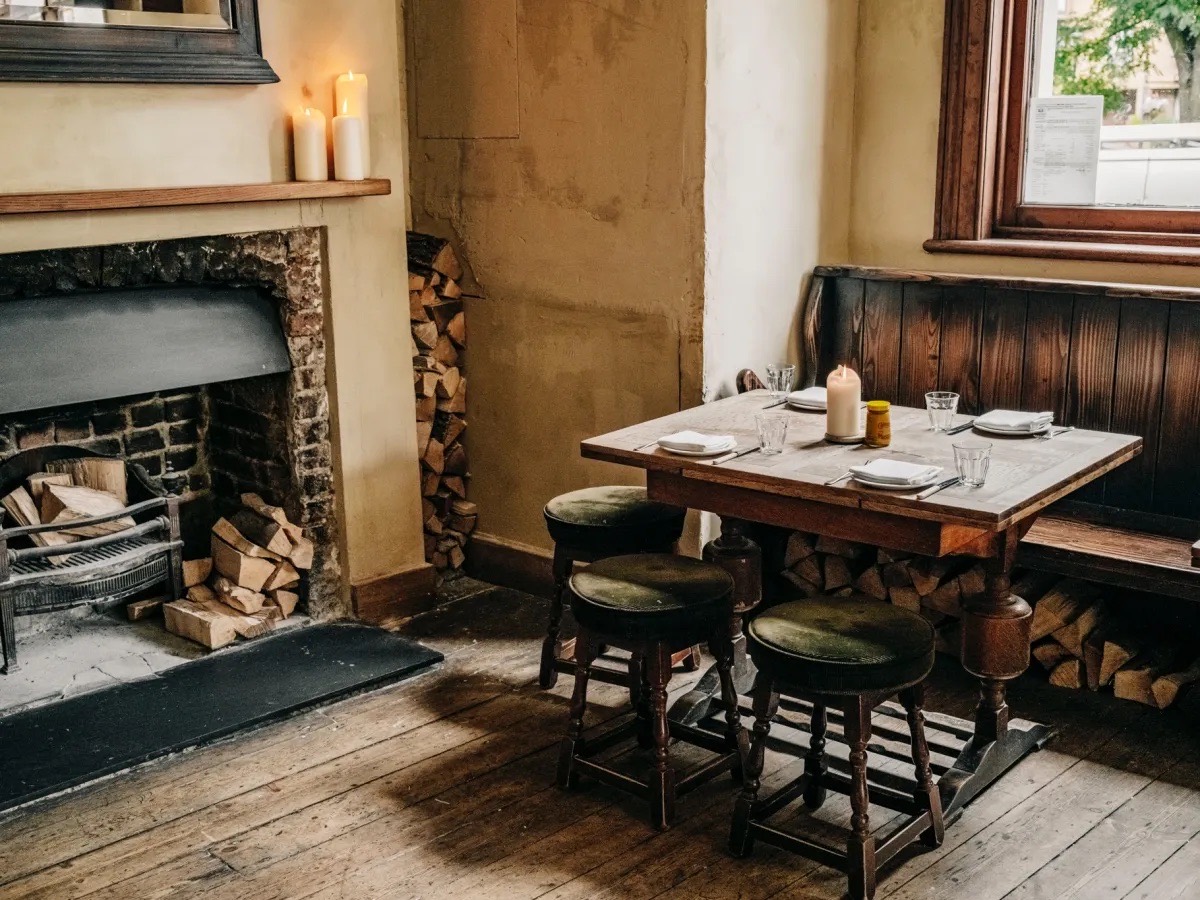 A pub corner table by a fireplace, logs stacked and candles lit